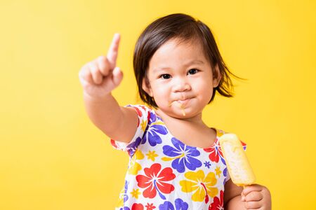 Happy portrait Asian baby or kid cute little girl attractive laugh smile wearing dick pattern shirt holds and eating sweet wooden ice cream, studio shot isolated on yellow background, summer conceptの写真素材