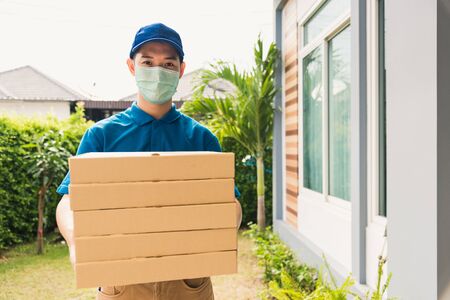 Asian young delivery man courier sending and holding fast food pizza boxed in uniform he protective face mask service customer at home door, under curfew quarantine pandemic coronavirus COVID-19の写真素材
