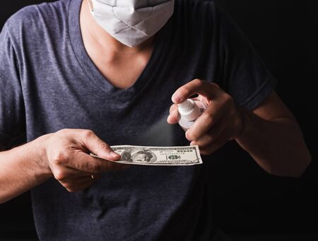 Closeup man wearing protective face mask applying sanitizer spray to 100 dollar money for cleaning,...