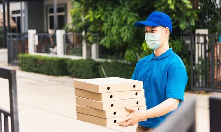 Asian young delivery man courier sending and holding fast food pizza boxed in uniform he protective face mask service customer at home door, under curfew quarantine pandemic coronavirus COVID-19の写真素材