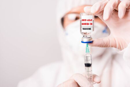 Asian female woman doctor or nurse in PPE uniform and gloves wearing face mask protective in lab hold needle syringe drug and medicine vial vaccine bottle on bottle has "COVID-19 VACCINE" text labelの写真素材
