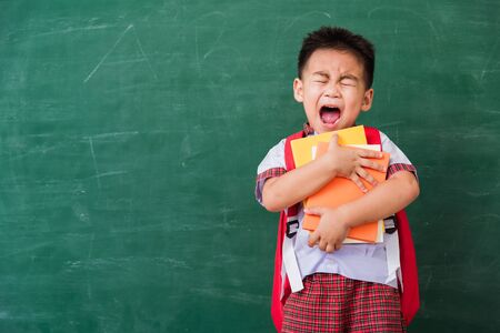 Back to School. Happy Asian funny cute little child boy from kindergarten in student uniform with school bag hold or hug books smile on green school blackboard, First time to school education conceptの写真素材