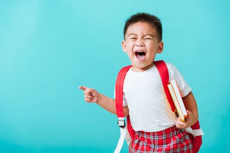 Back to school concept. Portrait Asian happy funny cute little child boy smile hug books and point finger to side away space, isolated blue background. Kid from preschool kindergarten with school bagの写真素材