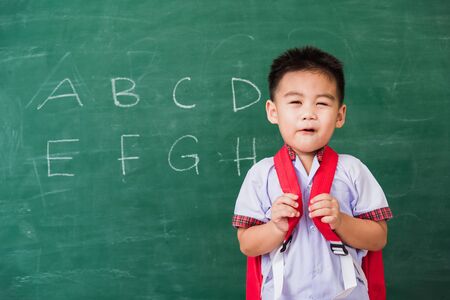 Back to School. Happy Asian funny cute little child boy kindergarten preschool in student uniform wearing school bag stand smiling on green school blackboard, First time to school education conceptの写真素材