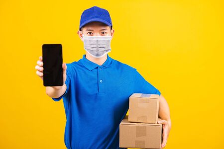Asian young delivery worker man t-shirt and cap uniform wearing face mask protective show front black screen of smart mobile phone under coronavirus or COVID-19, studio shot isolated yellow backgroundの写真素材