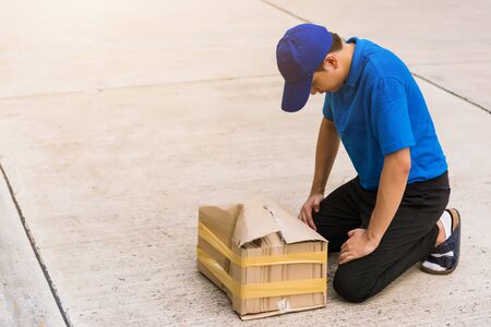 Asian young delivery man in blue uniform he emotional falling courier hold damaged cardboard box is broken at door front home, Accident bad transport shipment or poor quality delivery service conceptの写真素材