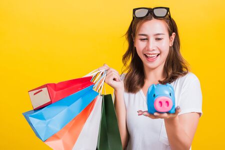 Portrait Asian Thai beautiful young woman happy hold colorful shopping bags and Piggybank Saving money, studio shot isolated on yellow background, with copy spaceの写真素材