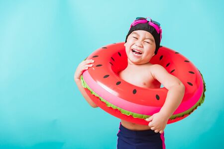 Summer vacation concept, Portrait Asian happy cute little child boy wear goggles and swimsuit hold watermelon inflatable ring, Kid having fun on summer vacation, studio shot isolated blue backgroundの写真素材