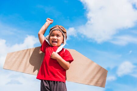 Happy Asian funny child or kid little boy smile wear pilot hat and goggles play toy cardboard airplane wing flying raises hand up against summer blue sky cloud background, Startup freedom conceptの写真素材