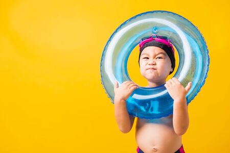 Summer vacation concept, Portrait Asian happy cute little child boy wear goggles and swimsuit hold blue inflatable ring, Kid having fun on summer vacation, studio shot isolated yellow backgroundの写真素材