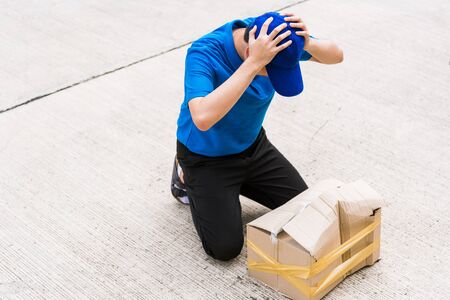 Asian young delivery man in blue uniform he emotional falling courier hold damaged cardboard box is broken at door front home, Accident bad transport shipment or poor quality delivery service conceptの写真素材