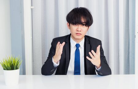 Work from home, Close up face of Asian young businessman video conference call or facetime he looking to camera raises hands to explain about his job to team on a desk at the home officeの写真素材