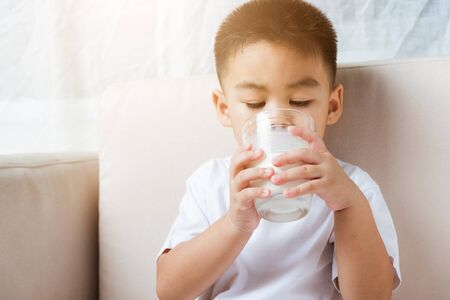 Close up of happy Asian little cute child boy hand holding milk glass he drinking white milk during sitting on the sofa at home after lunch. Daily life health care Medicine foodの写真素材
