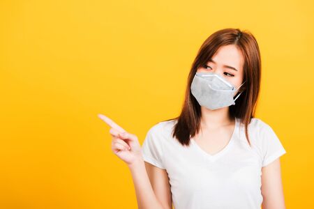 Portrait Asian beautiful happy young woman wearing face mask protects filter dust pm2.5 anti-pollution, anti-smog, and air pollution her pointing out to side on yellow background, with copy spaceの写真素材