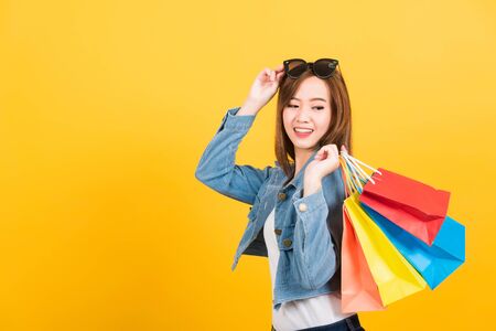Asian happy portrait beautiful cute young woman teen smiling standing with sunglasses excited holding shopping bags multi color looking bags isolated, studio shot yellow background with copy spaceの写真素材