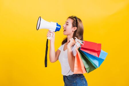 Portrait Asian Thai beautiful happy young woman smiling hold shopping bags multi-color and shouting in megaphone her looking to side away, studio shot isolated on yellow background, with copy spaceの写真素材