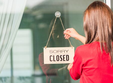 Asian young woman notice sign wood board label "SORRY WE ARE CLOSED PLEASE COME BACK AGAIN" hanging through glass door front shop, Business close during coronavirus pandemic disease conceptの写真素材