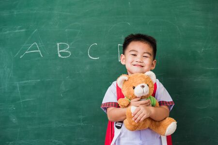 Back to School. Happy Asian funny cute little child boy from kindergarten in student uniform with school bag smiling and hugging teddy bear on green school blackboard, First time to school educationの写真素材