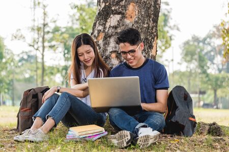 Young man and girls friend classmates sitting under tree consult on laptop at university togetherの写真素材