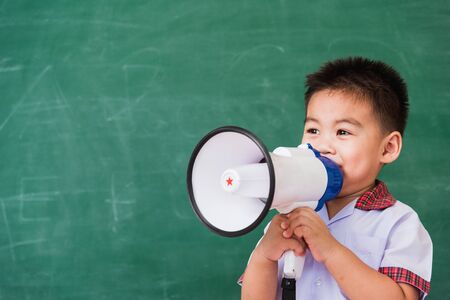 Back to School. Asian funny cute little child boy kindergarten preschool in student uniform speaking through megaphone against on green school blackboard, First time to school education conceptの写真素材