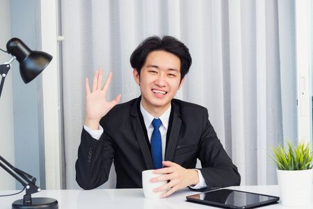Work from home, Asian young businessman video conference call or facetime he smiling looking to camera sitting on desk using smart digital tablet computer raise hand to say hello at home officeの写真素材