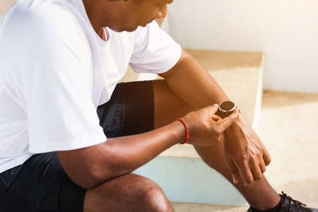 Close up Asian young athlete sport runner black man wear modern time smart watch he sitting resting before training running at the outdoor street health park, healthy exercise before workout conceptの写真素材