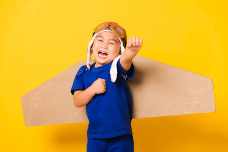 Happy Asian handsome funny child or kid little boy smile wear pilot hat play and goggles raise hand up with toy cardboard airplane wings flying, studio shot isolated yellow background, Startup freedomの写真素材