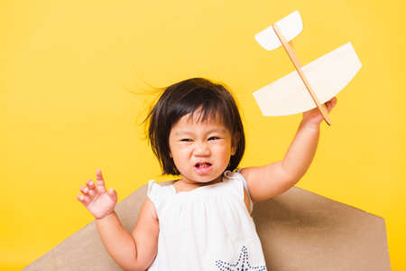 Happy Asian beautiful funny baby little girl smile wear pilot hat play with toy cardboard airplane wings fly hold plane toy, studio shot isolated yellow background, Startup freedom conceptの写真素材