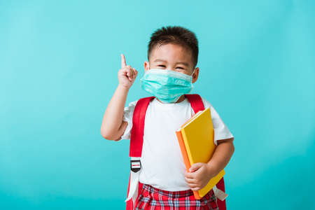 Back to school coronavirus Covid-19 education. Portrait Asian little child boy kindergarten wear face mask protective and school bag hold book before going to school pointing to side away, studio shotの写真素材