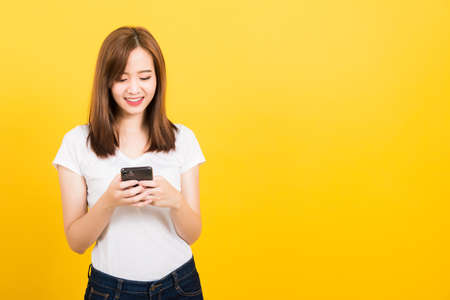Asian happy portrait beautiful cute young woman teen smile standing playing game or writing message on smartphone looking to the phone isolated, studio shot on yellow background with copy spaceの写真素材