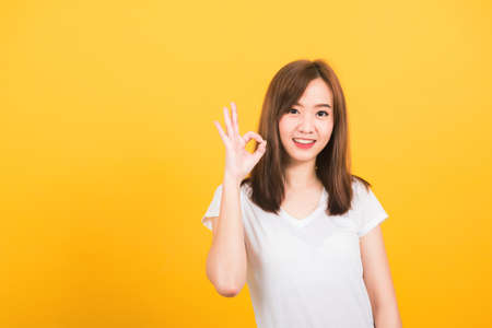 Asian happy portrait beautiful cute young woman teen standing wear t-shirt showing gesturing ok sign with fingers looking to camera isolated, studio shot on yellow background with copy space for textの写真素材