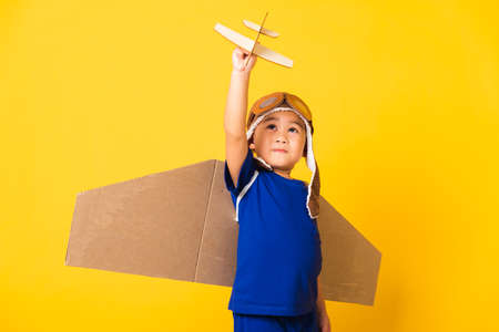 Happy Asian handsome funny child or kid little boy smile wear pilot hat play and goggles with toy cardboard airplane wings fly hold plane toy, studio shot isolated yellow background, Startup freedomの写真素材