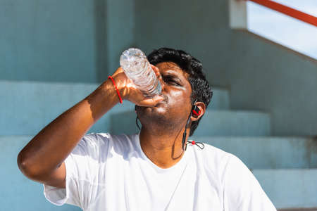 Close up Asian young sport runner black man wear athlete headphones he drinking water from a bottle after running at the outdoor street health park, healthy exercise workout conceptの写真素材
