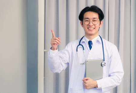 Portrait closeup of Happy Asian young doctor handsome man smiling in uniform and stethoscope neck strap holding smart digital tablet on hand and point finger to side away, healthcare medicine conceptの写真素材