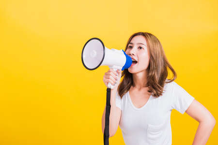 Asian Thai happy portrait beautiful cute young woman stand to make announcement message shouting screaming in megaphone looking to side away, studio shot isolated on yellow background with copy spaceの写真素材
