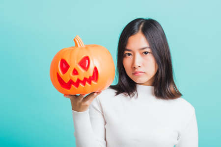 Portrait of Asian beautiful young woman holding orange model pumpkins, funny happy female with ghost pumpkins, studio shot isolated on blue backgroundの写真素材