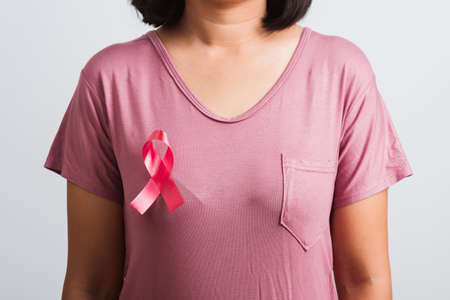 Breast cancer awareness healthcare and medicine concept. Close up Asian woman wear pink shirt standing with pink breast cancer awareness ribbon pin on chest, studio shot isolated on white backgroundの写真素材