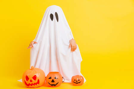 Funny Halloween Kid Concept, full body of a little cute child with white dressed costume halloween ghost scary he holding orange pumpkin ghost, studio shot yellow on white backgroundの写真素材