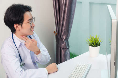 Asian young handsome doctor man wearing a doctor's dress and stethoscope video conference call or facetime raise hand to explain the symptoms he smiling at hospital office, Health medical care conceptの写真素材