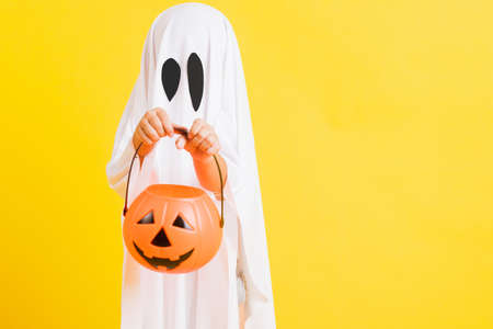 Funny Halloween Kid Concept, little cute child with white dressed costume halloween ghost scary he holding orange pumpkin ghost on hand, studio shot yellow on white backgroundの写真素材