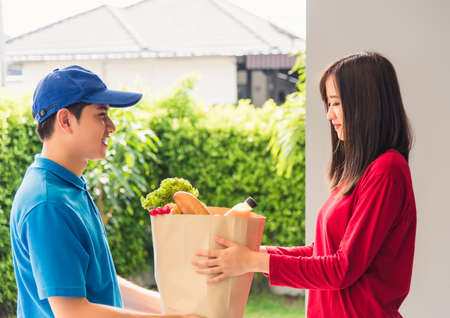 Asian young delivery man in uniform making grocery fast service giving fresh food in paper bag to...