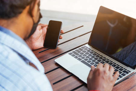 Happy Asian hand black man person working with laptop computer and reading message from modern smart phone blank screen device technology and connecting networking online at the coffee cafe shopの写真素材