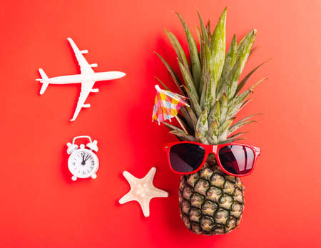 Summer Pineapple Day Concept; Top view flat lay of funny pineapple wear red sunglasses; model plane; starfish and clock alarm isolated on red background; Holiday summertime in tropicalの写真素材