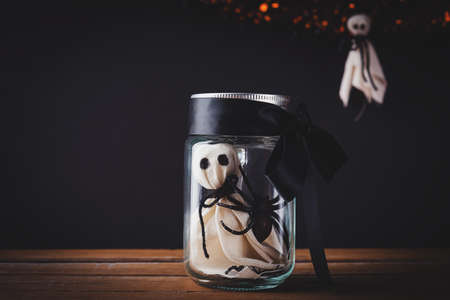 Funny Halloween day decoration party, The white ghost scary face and black spider in jar glass on wooden table, studio shot isolated on black background, Happy holiday conceptの写真素材