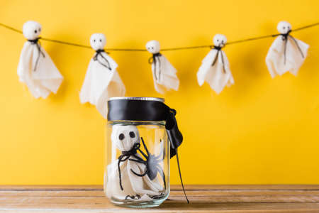Funny Halloween day decoration party, Cute white ghost crafts scary face hanging on background have only one baby ghost in jar glass, isolated yellow, Happy holiday DIY handicraft conceptの写真素材