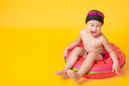 Summer vacation concept, Portrait Asian happy cute little child boy wear goggles, swimsuit hold beach watermelon inflatable ring, Kid have fun sit in inflatable, studio shot isolated yellow backgroundの写真素材