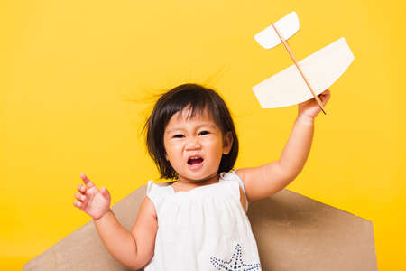 Happy Asian beautiful funny baby little girl smile wear pilot hat play with toy cardboard airplane wings fly hold plane toy, studio shot isolated yellow background, Startup freedom conceptの写真素材