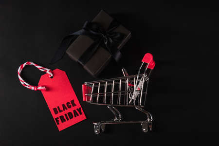 Black Friday sale shopping concept, Top view of gift box wrapped black paper and black bow ribbon present in the shopping cart and red tag label, studio shot on black backgroundの写真素材
