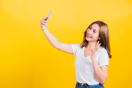 Asian Thai portrait happy beautiful young woman smiling making selfie photo or video call on smartphone she raise hand to say hello to smartphone, studio shot isolated yellow background and copy spaceの写真素材