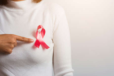 Breast cancer awareness healthcare and medicine concept. Close up Asian woman wear white shirt pointing finger to pink breast cancer awareness ribbon, studio shot isolated on white backgroundの写真素材
